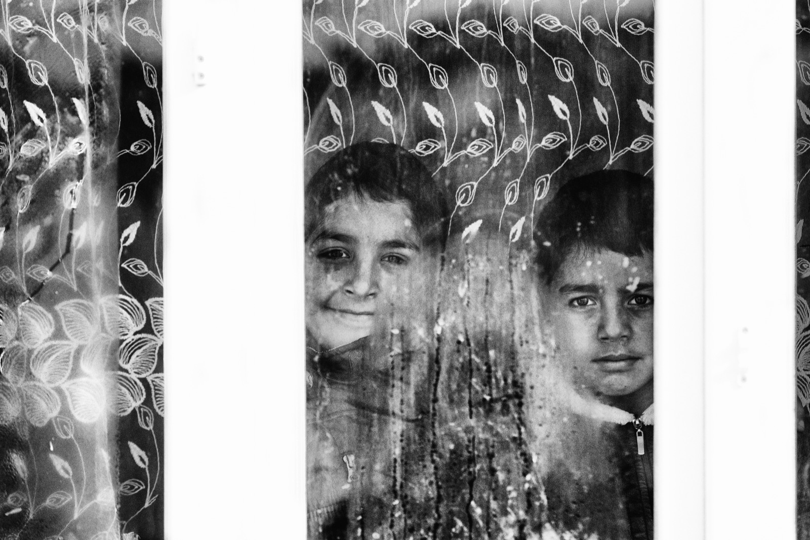 İki çocuk, kırağı ve dantel kaplı camın ardına sokulmuş; biri gülümsüyor, biri ciddi, Doğu Anadolu — Sefa Yamak'tan güzel sanatlar portre, En Zorlu Kış serisi.